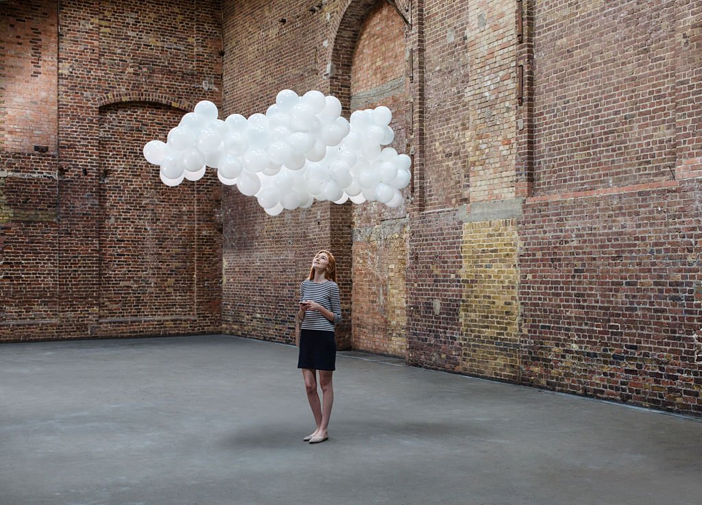 Young woman texting on mobile in empty warehouse with cloud made of balloons above head
